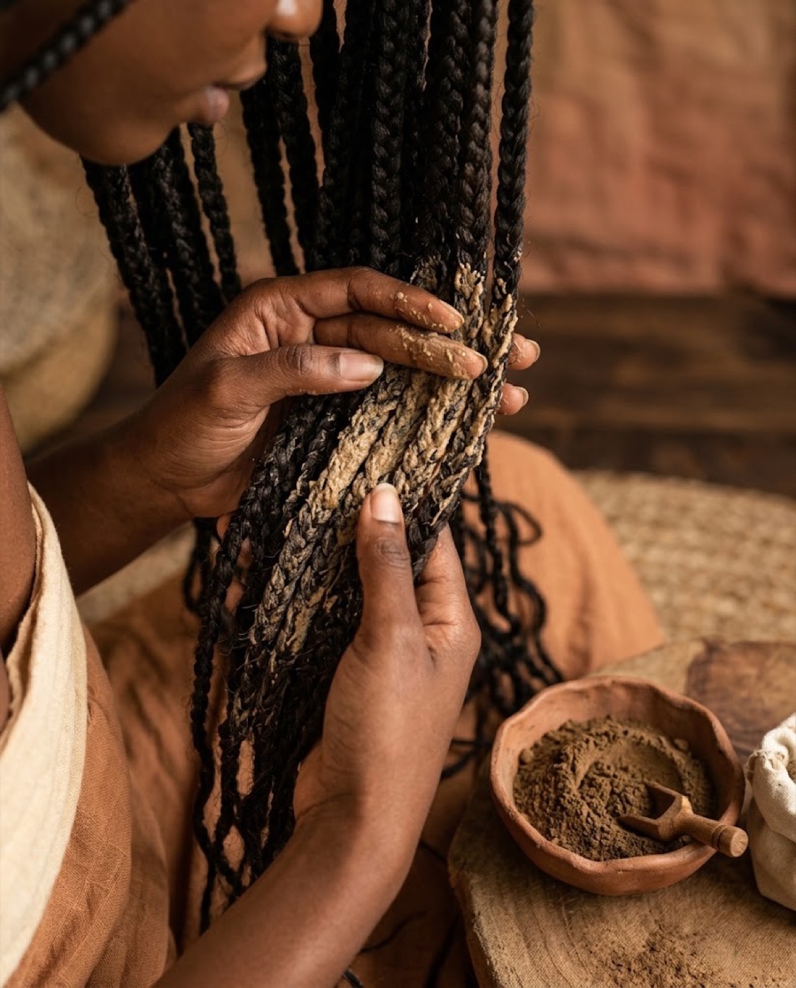 A woman applying a brown chebe paste to braided hair lengths, with a bowl of powder nearby