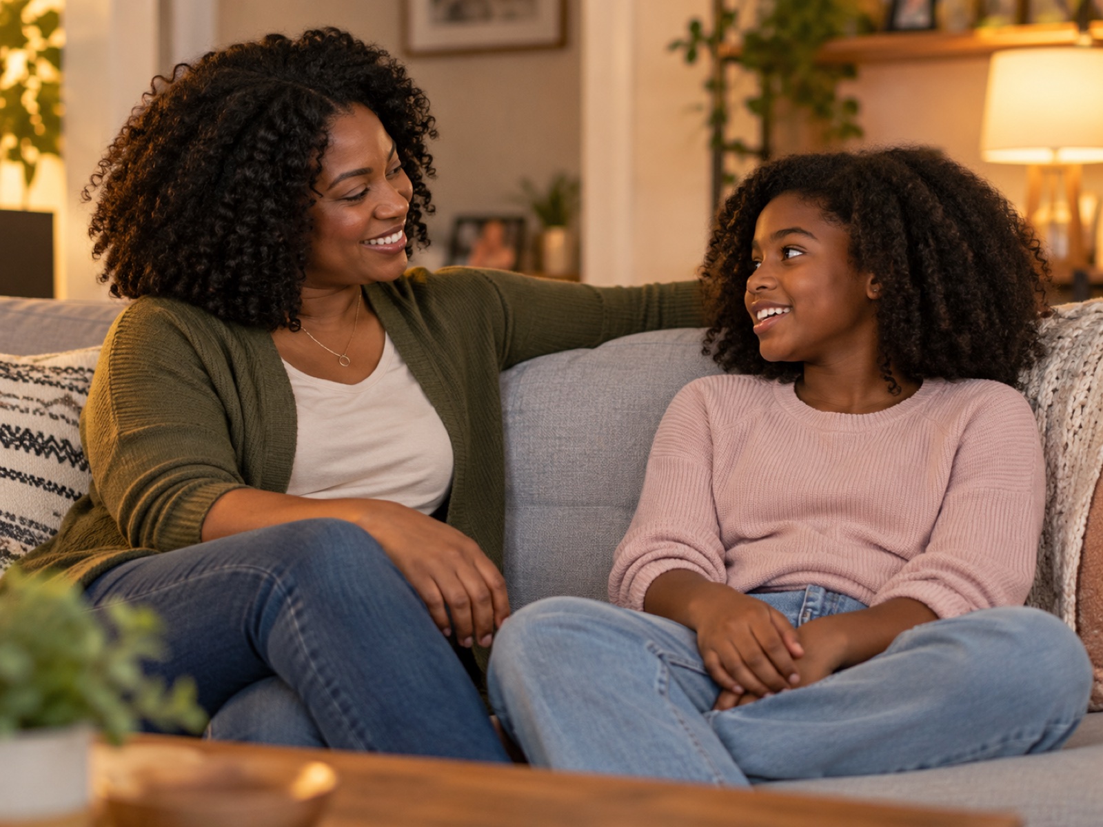 A Black mother and her preteen daughter talking and smiling on a living-room couch